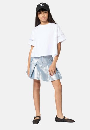 Chica con gorra de béisbol negra de Nueva York, camiseta blanca de gran tamaño, falda plisada plateada metálica y zapatos Mary Jane negros, de pie con la mano en la cadera.