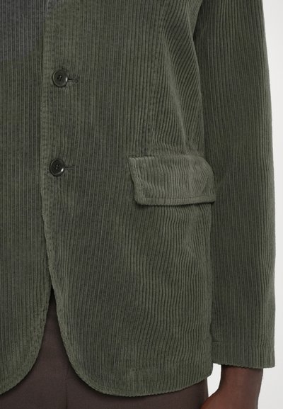 Jachetă din corduroy verde închis cu ribbing vertical, două butoane, un buzunar la piept și un tiv curbat. Material texturat cu o finisare moale.