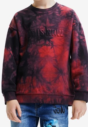 Dziecko ubrane w czerwoną i czarną bluzę typu tie-dye z napisem "JOY IS NOW" oraz niebieskie dżinsy z naszywką z kreskówką i napisem "NOW" na udzie.