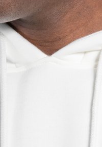 Sweat à capuche blanc en coton doux, avec un design en V, un tissu texturé et un cordon de serrage avec des accents tressés.