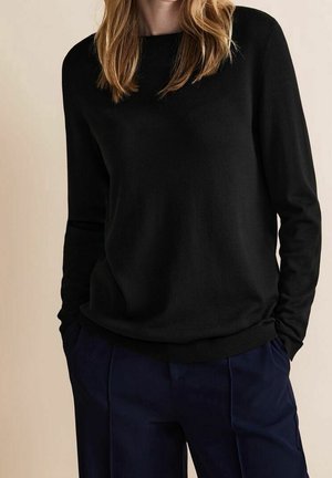 Personne portant un pull noir à manches longues et un pantalon bleu marine, les mains dans les poches, sur un fond beige.