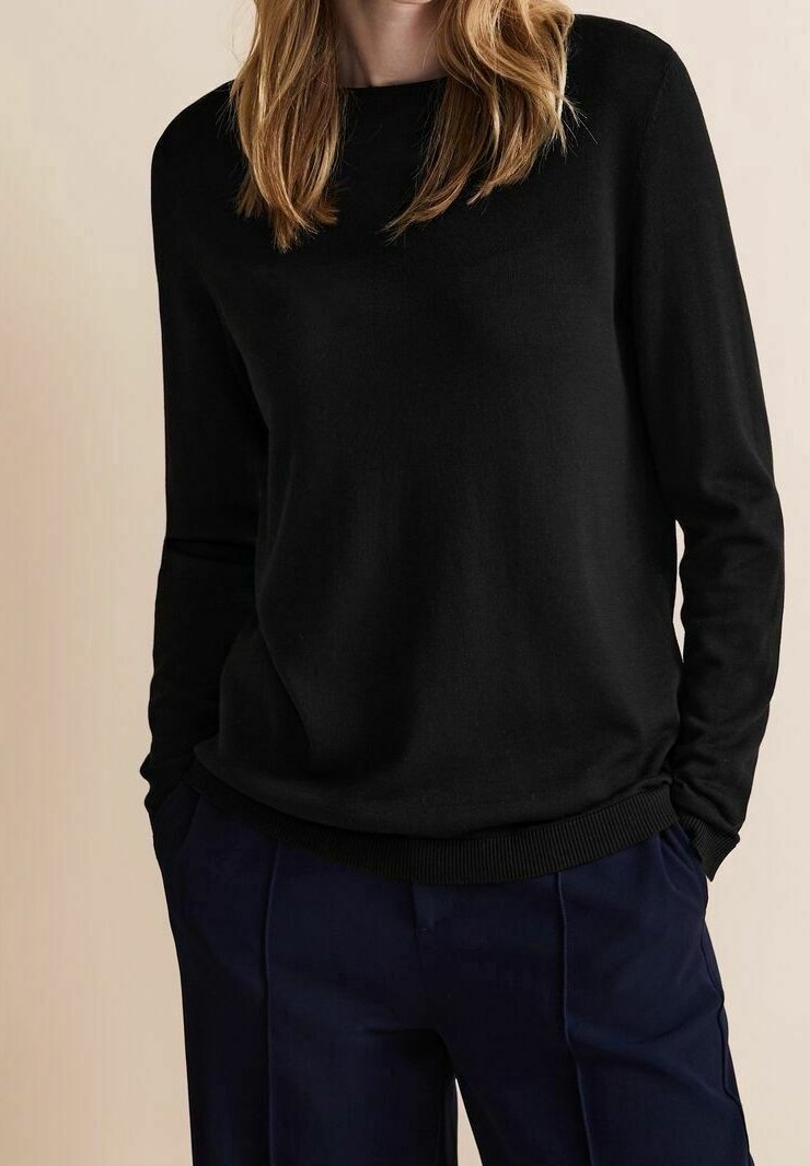 Personne portant un pull noir à manches longues et un pantalon bleu marine, les mains dans les poches, sur un fond beige.