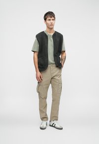 Un gilet en polaire gris sur un t-shirt vert, associé à un pantalon cargo beige. Le modèle se tient debout dans des baskets blanches avec des accents noirs devant un fond neutre.