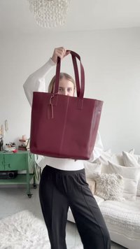 Sac fourre-tout en cuir bordeaux avec deux longues poignées, forme rectangulaire, texture lisse, et une étiquette discrète attachée, tenu par une personne.