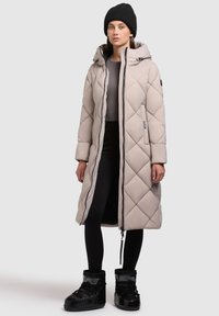 Lang, beige quiltet frakke med hætte, der har en frontlynlås, sidelommer og ribbede manchetter, parret med sorte støvler og leggings.