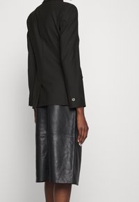 Blazer noir sur mesure avec épaules structurées et fermeture par un seul bouton, associé à une jupe en cuir longueur genou avec des détails de coutures.