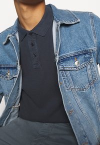 Veste en denim avec un délavage bleu clair et des boutons argentés, portée sur un polo bleu marine foncé avec un col et trois boutons.