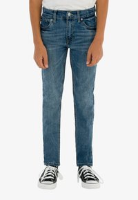 Jeans de denim azul de corte ajustado con un ligero desvanecimiento, que cuentan con cinco bolsillos, cierre de botón, y combinados con zapatillas negras con cordones blancos.