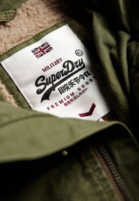 Zielona kurtka wojskowa w stylu, z beżowym, teksturowanym wykończeniem; na przodzie znajduje się biała i czerwona metka z napisem "SuperDry" oraz flagą Unii.