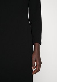 Robe noire ajustée présentant une texture côtelée et des manches trois-quarts, une main reposant sur le côté du vêtement.