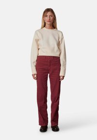 Off-white sweatshirt met lange mouwen, gecombineerd met bordeauxrode corduroy broek. Model draagt zwarte schoenen en staat op een witte achtergrond.