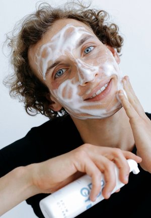 Flasche mit Gesichtsreiniger wird in einer Hand gehalten, mit Schaum im Gesicht. Weiße Flasche, simples Design, blauer Text. Heller Hintergrund.