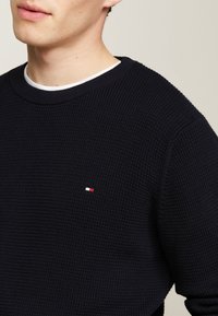 Maglione a maglia navy con un motivo testurizzato, dotato di un piccolo logo ricamato in rosso e bianco sul lato sinistro del petto. Girocollo.