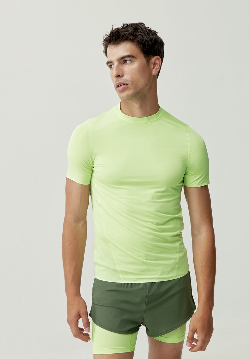 Maglietta atletica fitted verde chiaro a maniche corte con collo alto, realizzata in materiale liscio e stretchy, abbinata a pantaloni corti verdi scuri.