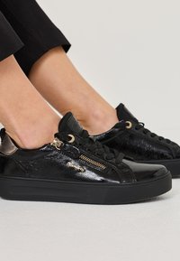 Fekete lakkbőr sportcipők texturált felülettel, arany színű részletekkel, fekete fűzőkkel és vastag fekete talppal. Oldalsó cipzár részlet található.