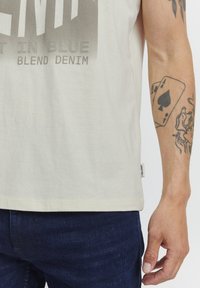 Camiseta de algodón en beige claro con un estampado gráfico en gris oscuro. Mangas cortas y corte recto, combinada con jeans azul oscuro.