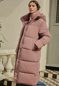 Palton lung, pufos, roz, cu guler înalt și glugă. Dispune de un fermoar în față, buzunare laterale și o textură matlasată. Modelul stă lângă un perete.