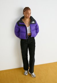 Lilla pufferjakke med svart krage, glidelås, kombinert med svarte bukser og grå sneakers. Enkelt design, teksturert stoff.