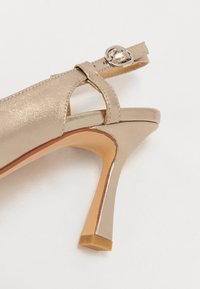 Sandalia de tacón alto de charol beige con diseño de tira en T y detalle de hebilla metálica. Presenta un tacón afilado y una textura suave.