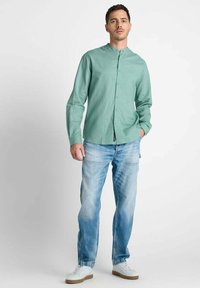 Man die staat met zijn handen in zijn zakken, gekleed in een groene knoopshirt, lichtblauwe spijkerbroek en witte sneakers tegen een effen achtergrond.