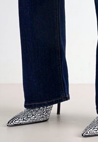 Talons hauts pointus argentés ornés avec un talon aiguille. Portés avec un pantalon en denim bleu foncé. Un motif en strass scintillant est bien visible.