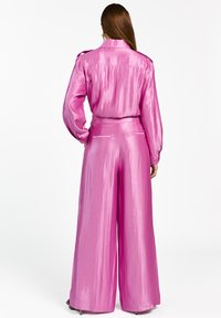 Vrouw met lang bruin haar, gekleed in een glanzende roze blouse met lange mouwen en wijde broek, staande met handen in de zakken, met haar rug naar ons toe.