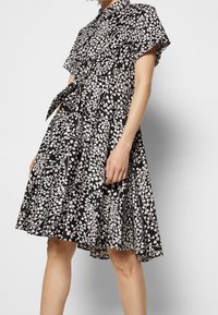 Schwarz-weiß gemustertes knielanges Kleid mit kurzen Ärmeln, Kragen und einem gebundenen Taillengürtel, getragen von einer stehenden Person vor einem schlichten Hintergrund.