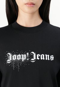 T-shirt en coton noir avec le texte blanc "joop! Jeans" présentant un effet dégradé de clous argentés, créant un détail texturé sur le devant.