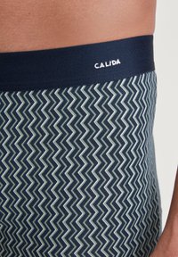 Navy blue patterned swim trunks with zigzag design in shades of green and white. Elastic waistband features logo "CALIDA." Soft, textured fabric.