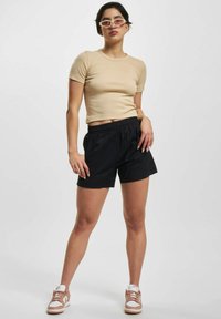 Ung kvinne iført beige crop top, svarte shorts, rosa og hvite joggesko, samt smale rosa solbriller, poserer foran en enkel hvit bakgrunn.