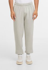 Persoon in lichtgrijze elastische sweatpants en witte sneakers, staande tegen een effen witte achtergrond.