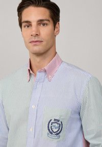 Hombre que lleva una camisa de botones de rayas verticales multicolores con un cuello a rayas rojas y un emblema bordado en el bolsillo del pecho.