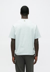 Szara bawełniana koszulka z krótkimi rękawami, luźnym fasonem i małym zielonym logo na karku. Gładka tekstura z okrągłym dekoltem.