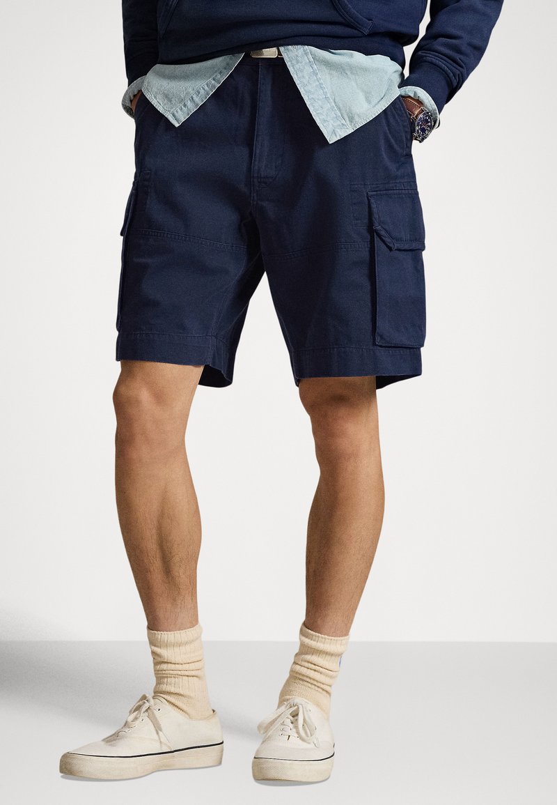 Tumši zili kokvilnas šorti ar sānu kabatām, kopā ar gaiši zilu džinsa kreklu un krēmkrāsas sporta apaviem ar ribotiem bēšiem zeķēm.