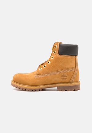 Brown leather ankle boot with black padding, gold eyelets, and a textured sole. Features a circular logo on the side and stitching accents.