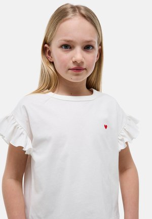 Jeune fille aux longs cheveux blonds portant une chemise blanche à manches froufroutantes avec un petit cœur rouge brodé sur la poitrine.