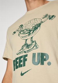 Beige bomulds t-shirt med et grønt grafik af en hamburger, der løfter vægte, med teksten "BEEF UP" nedenunder og Nike-logo på siden.