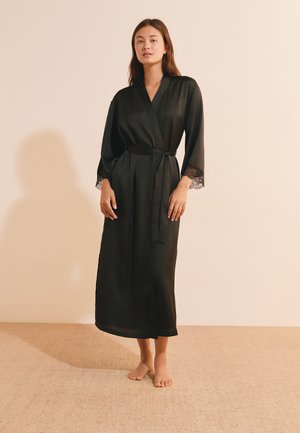Femme debout pieds nus portant une longue robe en satin noire avec des bordures en dentelle aux manches, nouée à la taille sur un fond beige uni.