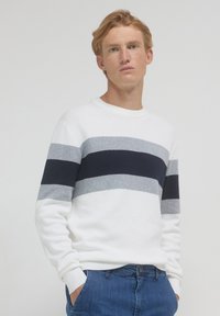 Giovane uomo con i capelli rosso chiaro, indossa un maglione bianco con strisce orizzontali grigie e nere e jeans blu, in piedi davanti a uno sfondo semplice.