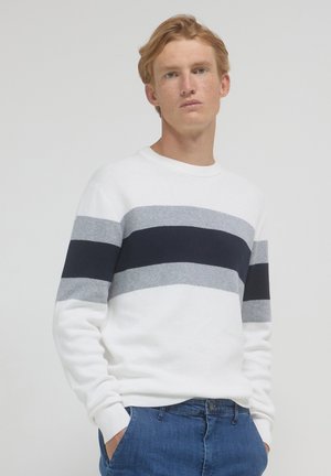 Junger Mann mit hellrotem Haar, der einen weißen Pullover mit grauen und schwarzen horizontalen Streifen und blaue Jeans trägt, steht vor einem schlichten Hintergrund.