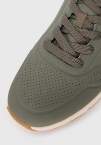 Adidași verde olive cu un superior din cauciuc neted, perforații texturate și șireturi plate maro. Prezintă o talpă de culoare deschisă cu un vârf rotunjit.