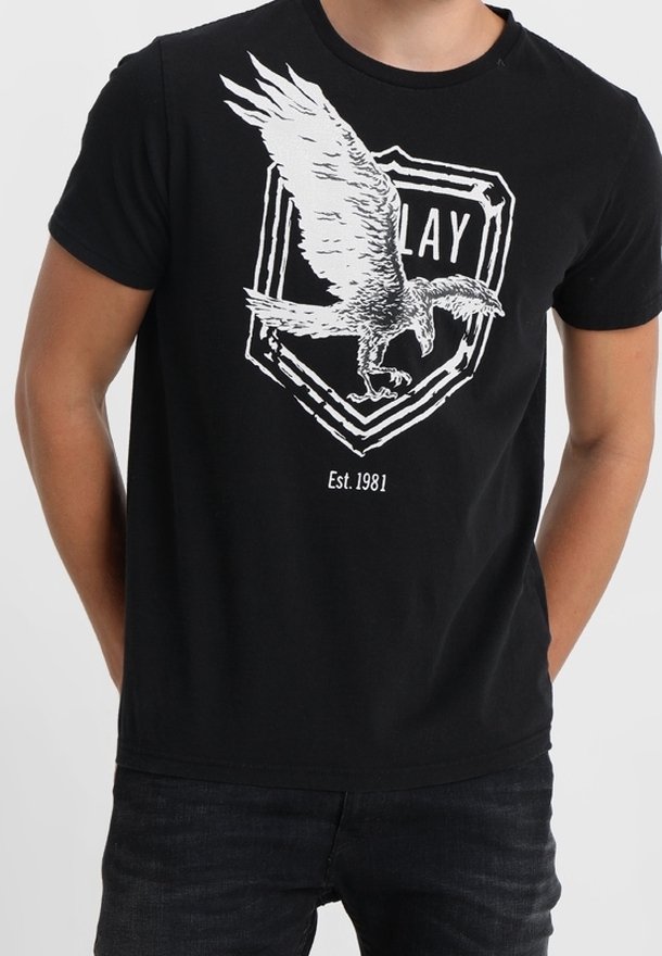 T-shirt noir avec un graphisme blanc représentant un grand aigle en vol au-dessus d'un bouclier, avec les textes "PLAY" et "Est. 1981" sur le devant.