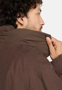 Giacca marrone con colletto rialzato, caratterizzata da un tessuto testurizzato e dettagli in Velcro. Il colletto viene regolato con una mano.