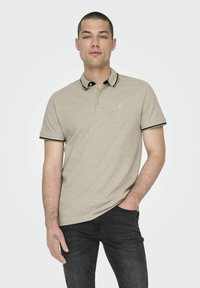 Beige poloshirt met zwarte en groene accenten op de kraag en mouwen. Glad textuur, korte mouwen en minimale logo-details.