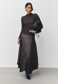 Haut à manches longues texturé marron associé à une maxi jupe satinée marron foncé et un sac seau en cuir noir. Le mannequin porte des bottines noires pointues.