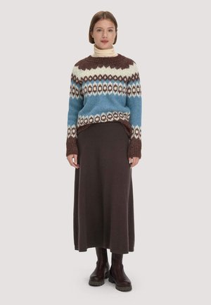 Frau trägt einen blau-weiß-braun gemusterten Pullover über einem beigen Rollkragenpullover, einen dunkelbraunen langen Rock und dunkelbraune Stiefel, steht aufrecht.