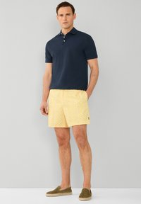 Mornarska polo majica, kratke hlače s žutim uzorkom i maslinaste slip-on cipele. Hlače imaju geometrijski uzorak. Glatka tekstura; opušteni kroj.