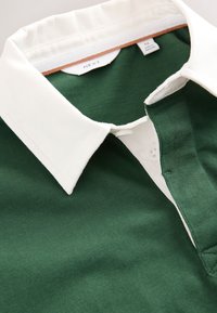 Polo vert avec col blanc, en coton texturé. Dispose d'une patte de boutonnage à deux boutons et d'une étiquette de marque à l'intérieur du col.