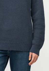 Άτομο που φοράει σκούρο μπλε πλεκτό πουλόβερ με ριμπ μανσέτες και τελείωμα, συνδυασμένο με μπλε τζιν, στέκεται μπροστά σε απλό φόντο.