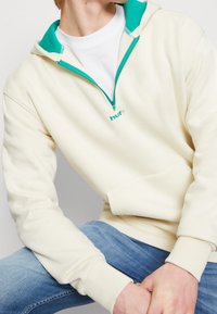 Sweat à capuche crème zippé avec doublure intérieure sarcelle, arborant un petit logo sur la poitrine et des poignets côtelés. Associé à un jean en denim bleu.